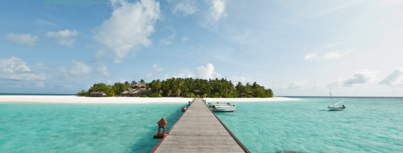 Segeln auf den Malediven, Steg inmitten türkisblaues Meer mit Ausblick auf die grüne Insel, Boote auf dem Wasser