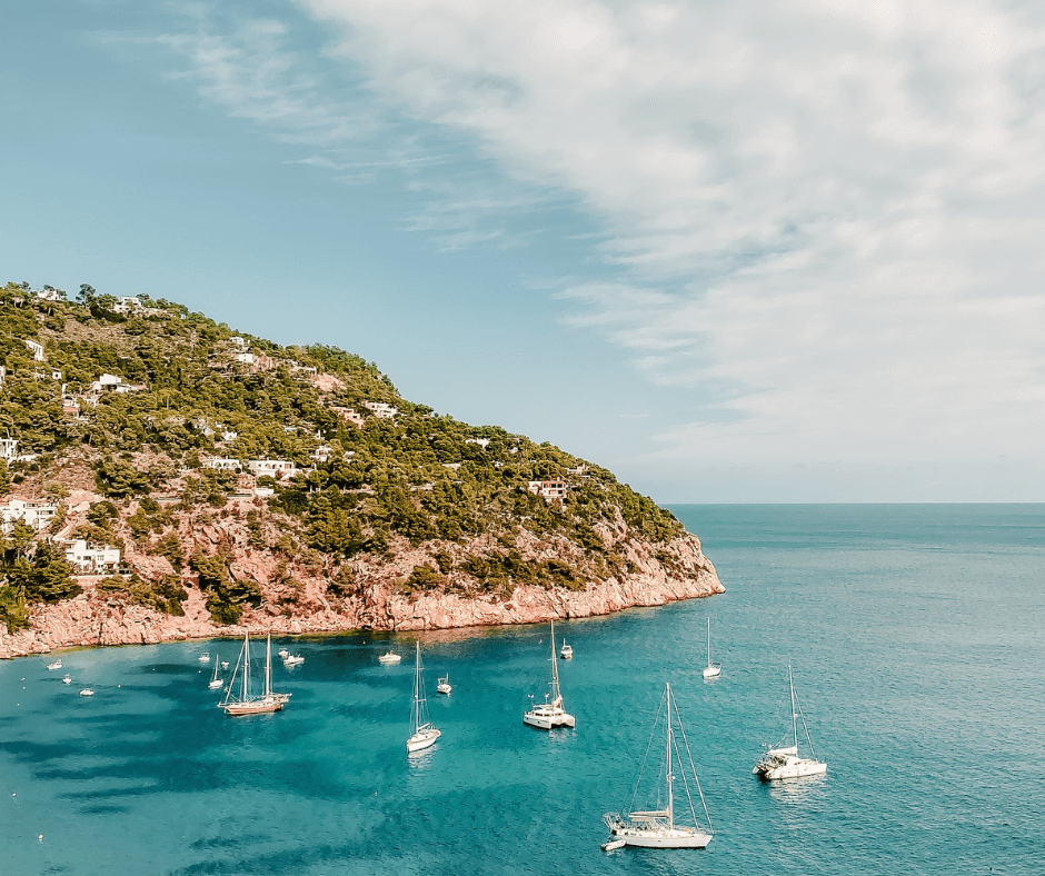 Verschiedene weiße Boote vor der grün bewachsenen Küste Ibizas, die im blauen Wasser liegen