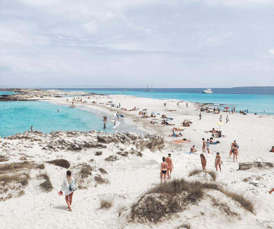 Strand auf Formentera mit weißem Sand, blauem Wasser, Booten am Horizont und verschiedenen Menschen, die baden oder sich am Strand sonnen