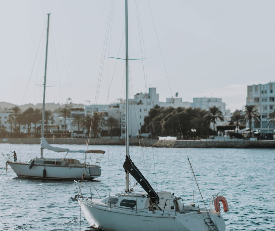Zwei Segelboote vor der Küste Ibizas mit Häusern und Palmen im Hintergrund