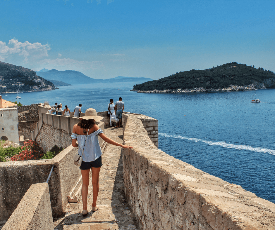 Spaziergänger und Spaziergängerinnen auf der Stadtmauer rund um Dubrovnik mit Blick auf die davorliegende Insel und die bergige Küste bei sonnigem Wetter