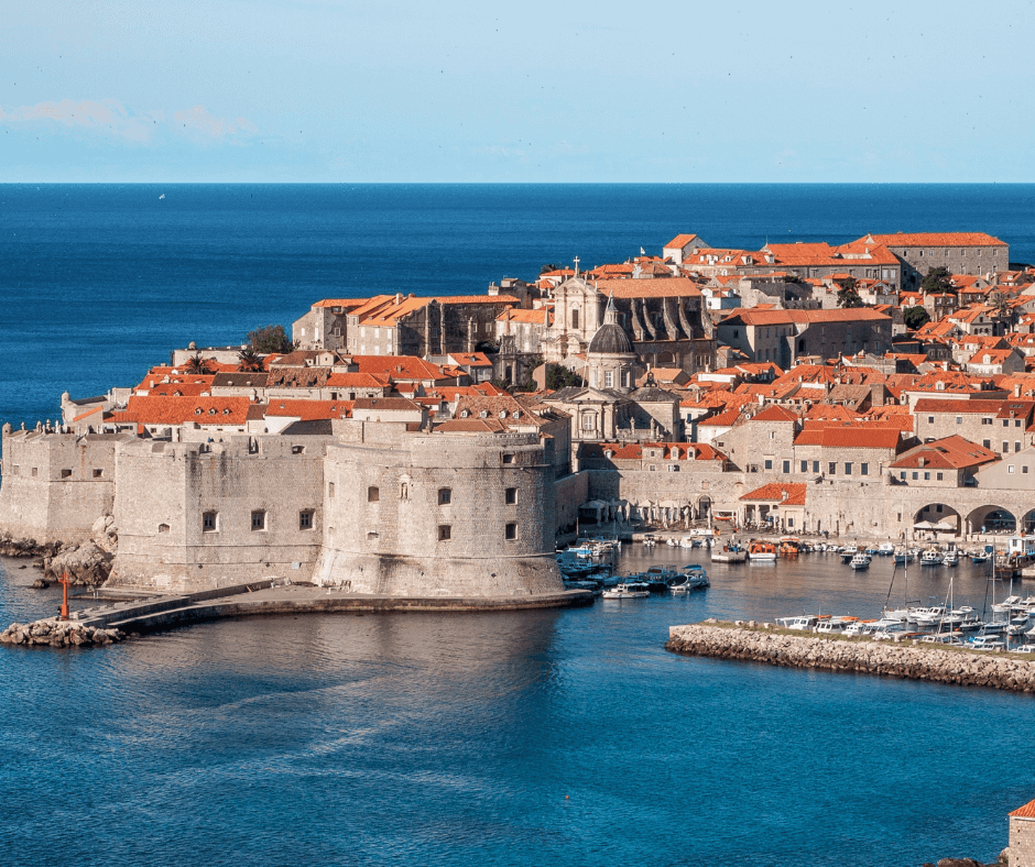 Burg und historische Altstadt von Dubrovnik aus der Vogelperspektive aufgenommen. Die Stadt ist umgeben von blauem Meer. Im Vordergrund ist der kleine Hafen. 