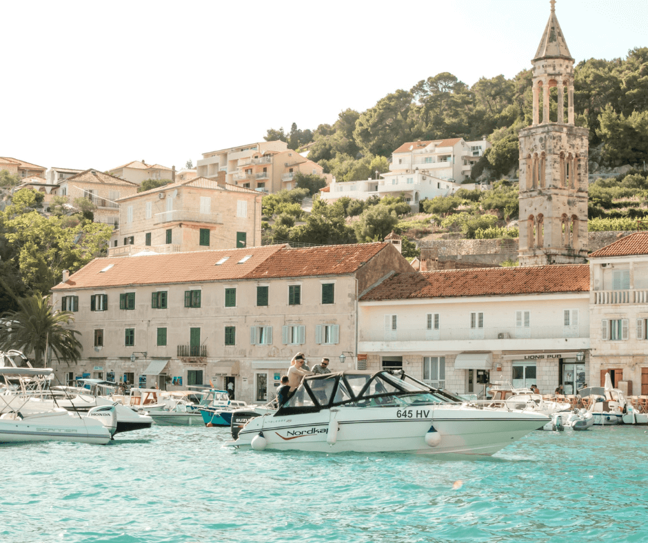 Inselhopping Kroatien - Insel Hvar, Hafen von Hvar, Häuser und alter Turm am Ufer, Motorboote mit Personen auf dem türkisfarbenen Wasser, grüne Landschaft im Hintergrund