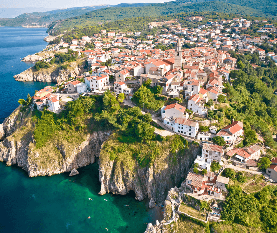 Inselhopping Kroatien - Insel Krk, Stadt, weiße Häuser mit orangen Dächern, Kirchenturm, grünbewachsene Hügellandschaft inmitten türkisblaues Wasser, Personen schwimmen neben einer Klippe