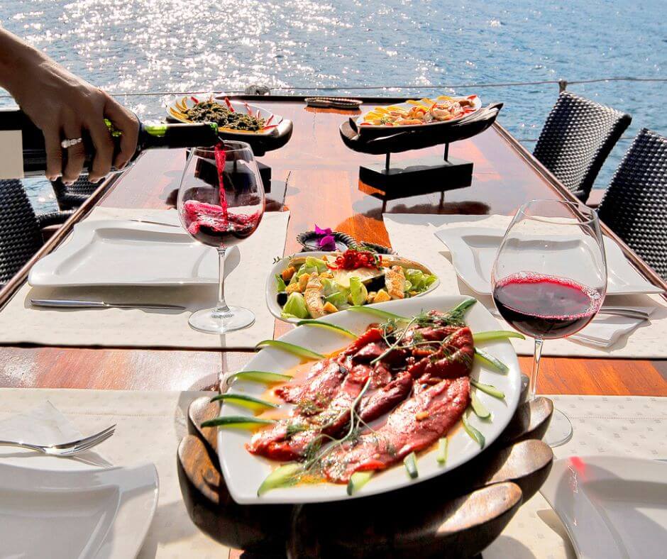 Gedeckter Tisch mit Tellern voller Essen und zwei Weinglasern. Ein Weinglas wird eingeschenkt, während das andere voll ist. Im Hintergrund sieht man das Meer.
