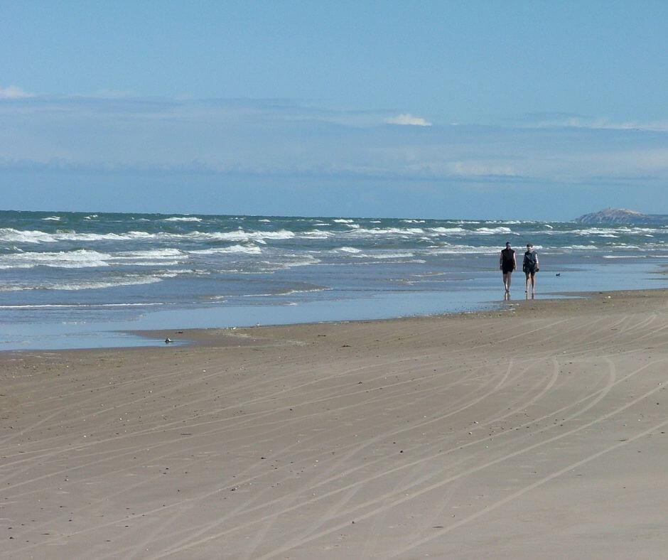Zwei Menschen, die einen Spaziergang am Sandstrand machen. Im Hintergrund ist das Meer zu sehen.