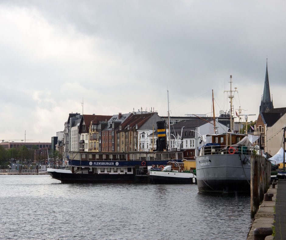 Ausschnitt des Hafens von Flensburg. Auf dem Wasser befinden sich 2 Passagierboote und ein kleines Fischerboot. Rechts davon ist eine kleine Promenade mit bunten Wohnhäusern. 