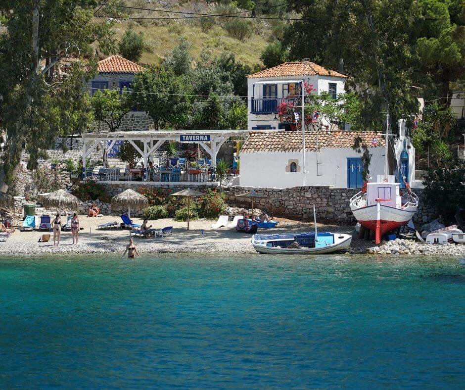 Türkises Wasser und ein kleiner Strand, an dem Menschen, Boote und Sonnenschirme zu sehen sind. Dahinter steht ein weißes Haus.