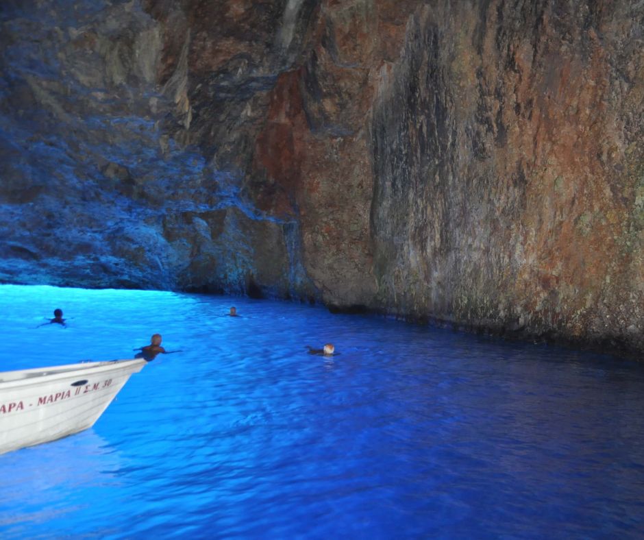 Meerwasser in einer Höhle, das in verschiedenen Blautönen schimmert. Menschen schwimmen im Meer und man sieht die Spitze eines kleinen Bootes.