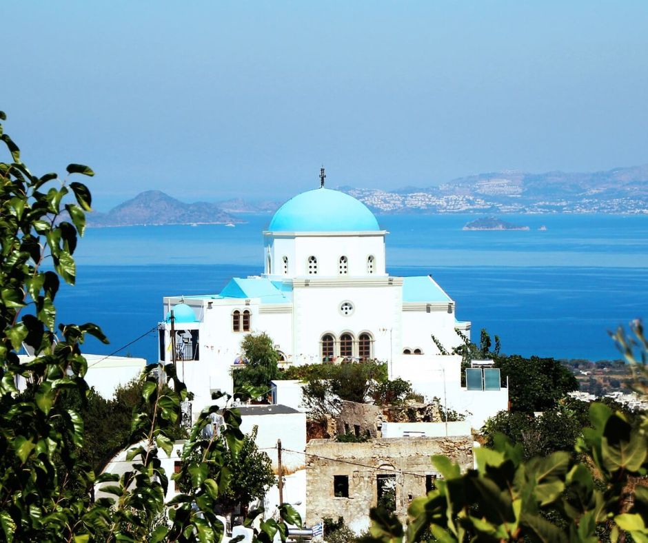 Kirche auf Kos im Vordergrund. Die Kirche hat ein blaues Dach und ist weiß. Im Hintergrund sieht man eine Aussicht auf das Meer mit anderen Inseln.