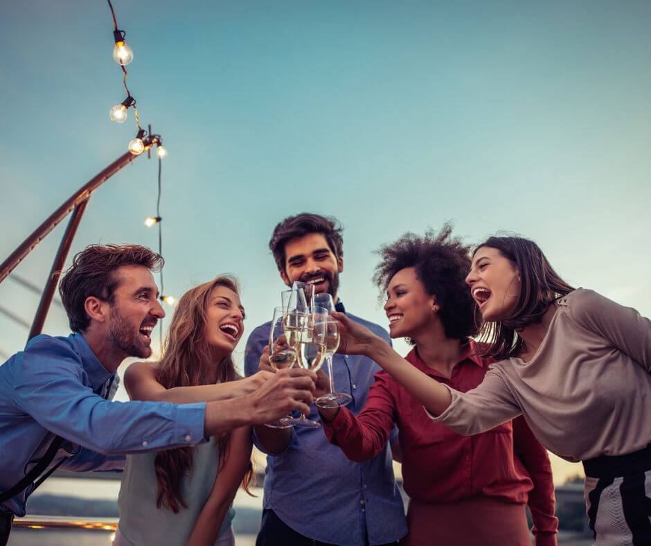 Fünf Personen, die mit Sekt anstoßen. Links im Bild sieht man eine Lichterkette und den blauen Himmel.