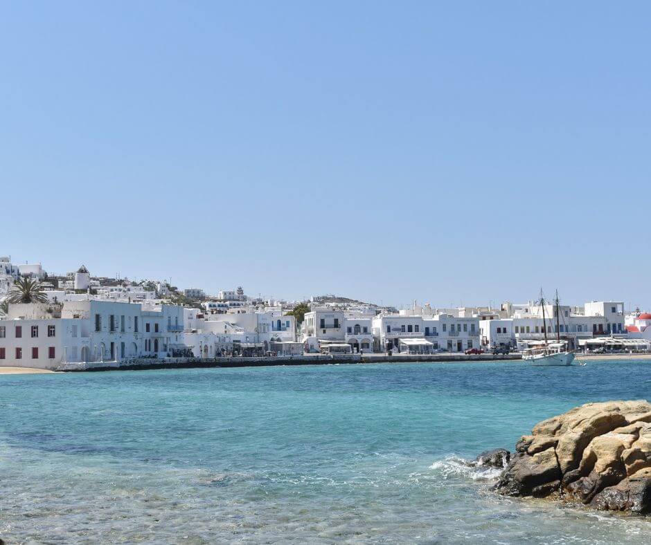 Promenade von Mykonos mit weißen Häusern. Ein Boot liegt auf dem Wasser. Vorne rechts sieht man einen Felsen.