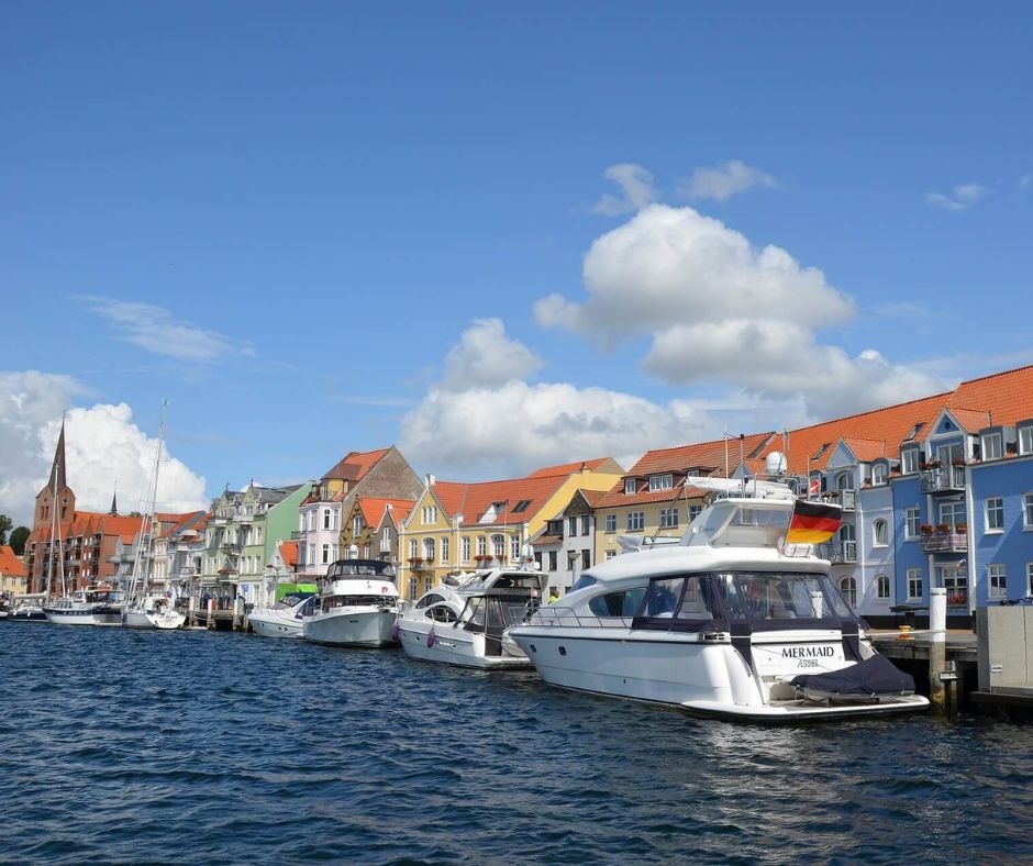Blick vom Wasser auf die Promenade von Sonderborg, mit bunten Häusern im Hintergrund. An der Seite ankern vreschiedene Yachten und Segelboote.