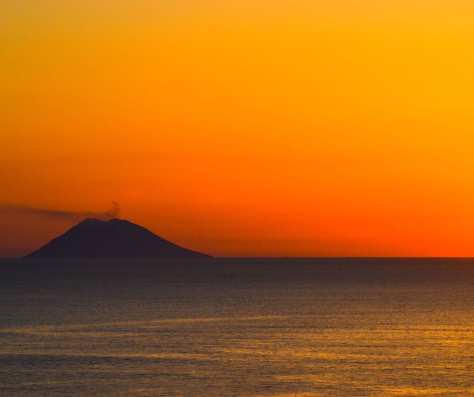 Insel mit Vulkan Stromboli bei Sonnenuntergang mit Meer im Vordergrund.