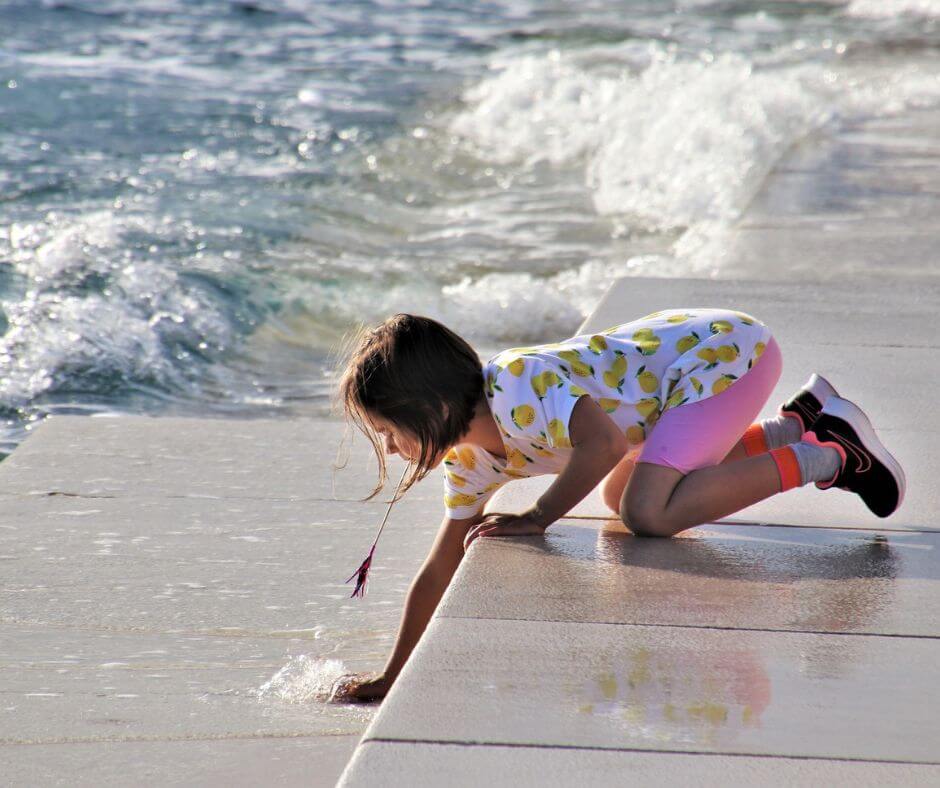 Ein Mädchen am Wasser von Zadar. Sie kniet und streckt eine Hand in das Wasser.