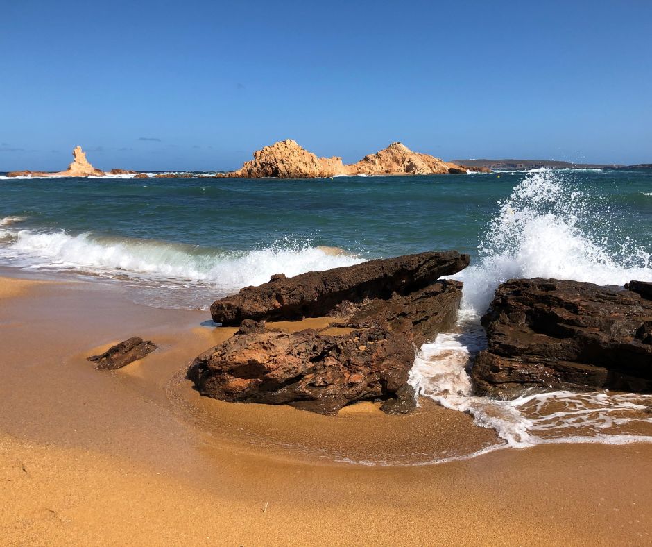 Goldbrauner Sand mit tiefblauem Meer im Hintergrund, das sich an Felsen im Meer bricht. Im Hintergrund befindet sich ein Felsmassiv im Wasser.